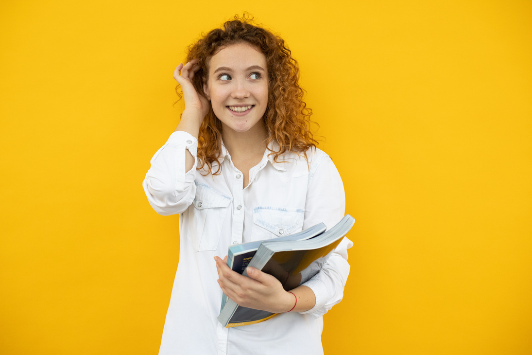 Cheerful female student with textbooks touching hair in studio